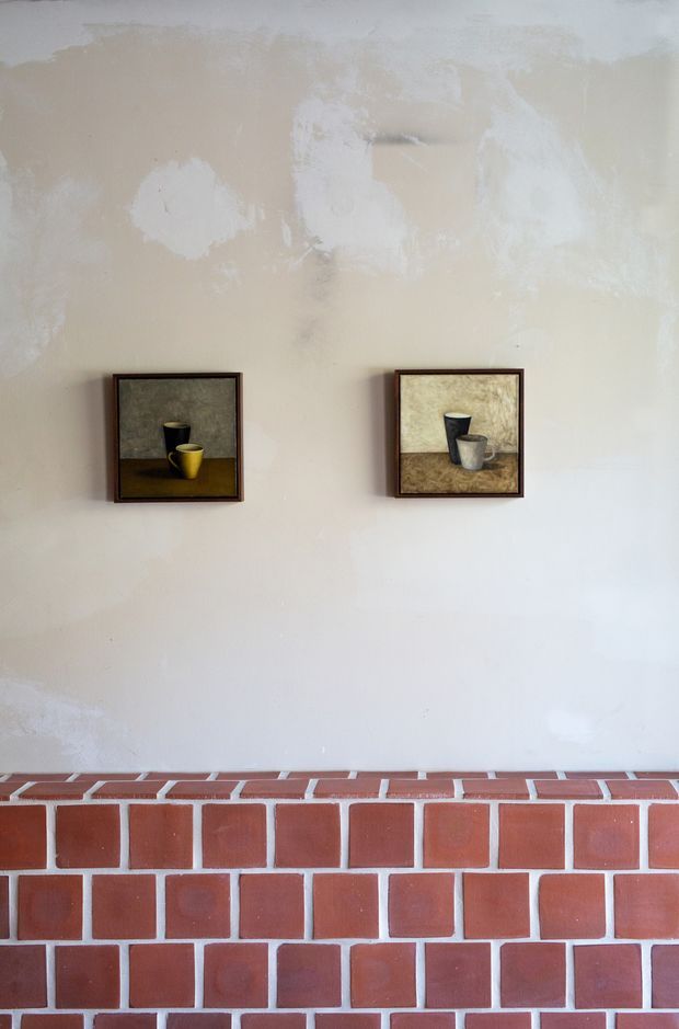 A close-up interior view of a textured pale plaster wall above a red brick fireplace or hearth. Two small framed still-life artworks depicting cups are symmetrically hung on the wall. The scene has a simple, warm, rustic character with visible brickwork and softly irregular wall finish.