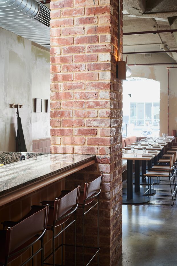 Interior view of an industrial-style restaurant or hospitality venue, with a prominent exposed red-brick structural column dividing a polished stone bar counter and leather bar stools from rows of neatly set dining tables. The space features exposed concrete ceilings, visible mechanical ductwork and services, dark metal furniture, large glazing, and a polished concrete floor.