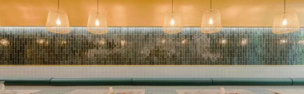 A close-up view of a refined commercial interior feature wall. The lower wall is clad in small pale rectangular tiles with a dark green upholstered or padded horizontal bench running along it. Above, a reflective mosaic tile backsplash in muted green, grey, and bronze tones extends beneath a warm tan ceiling. A row of woven pendant lights creates a warm, softly illuminated atmosphere.