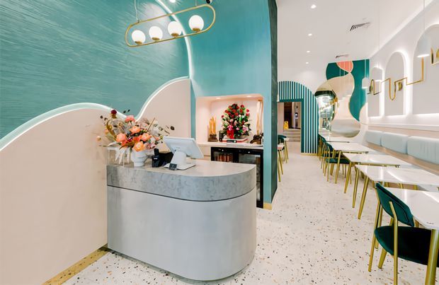 A brightly styled contemporary commercial interior, likely a café or hospitality venue, viewed from the front reception or service counter. The space features a curved light-grey stone or concrete counter, terrazzo flooring, a textured teal feature wall, arched white wall detailing, banquette seating, small white tables, teal-and-gold chairs, decorative pendant lighting, and a floral display. The overall design is playful, refined, and highly graphic, with teal, white, pale pink, and brass-gold accents.