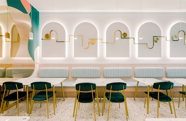 A brightly lit contemporary commercial dining space with a row of small tables and dark green upholstered chairs with slender brass-coloured legs. The back wall features repeated arched recessed niches with integrated halo lighting, pale upholstered banquette seating, and playful multicoloured linear wall detailing. A curved decorative wall element and teal, cream, and mustard accents add a bold, refined character. The terrazzo floor and coordinated custom joinery reinforce the polished hospitality interior.