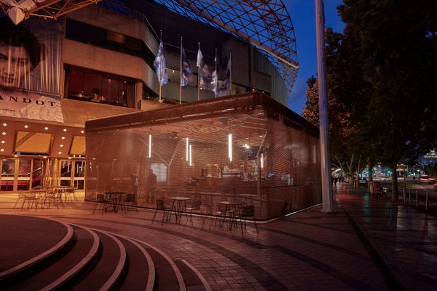 Dusk view of a contemporary commercial building and street-facing outdoor dining terrace. The foreground features curved stepped paving and an enclosed terrace with fine metal mesh walls, café tables and chairs, warm interior lighting, and a large overhead steel canopy structure. Urban streetscape and mature trees are visible to the right.