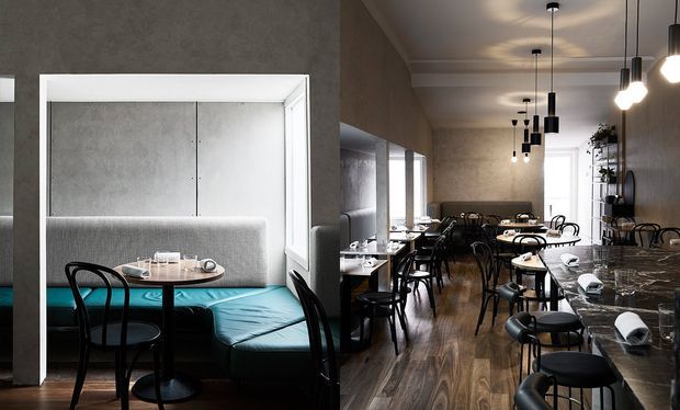 A long, narrow contemporary café or restaurant interior with dark timber-look flooring, marble-topped tables, black bentwood-style chairs, and a row of black pendant lights. A striking built-in booth with a turquoise upholstered bench and pale grey textured backrest occupies the foreground, while additional dining seating and wall shelving extend toward the rear.