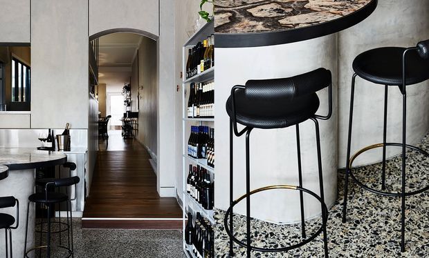 A stylish contemporary kitchen and bar area with a curved raised bar counter, black sculptural bar stools, open bottle shelving, and speckled terrazzo flooring. A narrow hallway with dark timber flooring extends through the centre of the image, while pale textured walls and dark accents create a refined, monochromatic interior.