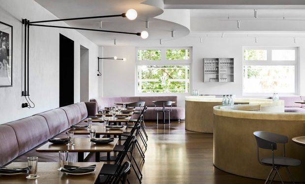 A bright, spacious contemporary restaurant interior with a long upholstered banquette and rows of small dining tables and black chairs along the left wall. Curved cream-toned built-in seating and a rounded service counter occupy the right side, while large windows, white walls, polished timber flooring, and exposed track-style lighting create an airy, refined atmosphere.