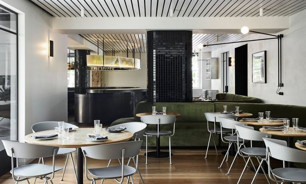 A bright contemporary restaurant or hospitality dining interior with multiple round and rectangular timber dining tables, white modern chairs, dark green upholstered banquette seating, and a black tiled central column. The space features a warm timber floor, white walls, a striking linear slatted ceiling, black-framed glazing, pendant lighting, and a dark bar or service counter in the background.