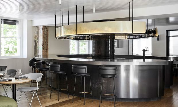 A sophisticated contemporary commercial bar or hospitality interior dominated by a large curved stainless-steel bar counter with black stools. A dramatic oversized suspended oval light fixture hangs above the bar, while black tiled detailing, reflective metal surfaces, exposed textured masonry, timber flooring, and a small dining area create a polished industrial atmosphere. Large windows provide natural light, and a service tap and back-bar area are visible.