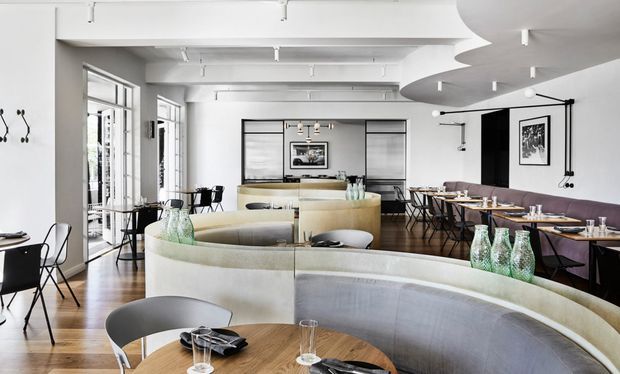 Bright contemporary restaurant interior with a large curved upholstered banquette surrounding round timber dining tables in the foreground. Additional dining tables and chairs occupy the open-plan space, with a central curved service counter, full-height windows, white exposed ceiling beams, track lighting, and a dark accent wall with framed artwork. The space has a refined, modern hospitality fit-out with warm timber flooring and neutral upholstery.