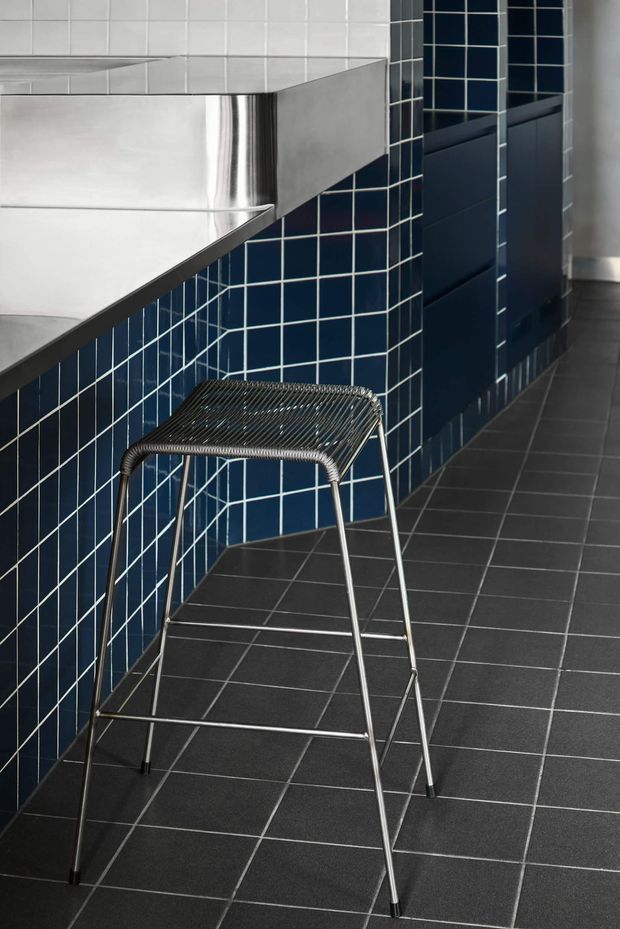 Close-up architectural interior detail showing a dark navy tiled wall and matching tiled floor, with a stainless-steel counter or ledge projecting from the wall. A minimalist metal-framed woven bar stool stands in the foreground. The composition is strongly geometric, with glossy blue square tiles, pale grout lines, and reflective stainless steel.