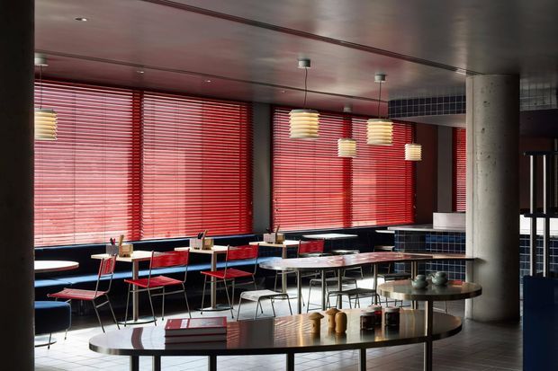 A stylish contemporary dining and lounge area with long communal tables, red dining chairs, a dark curved table in the foreground, pendant lights, and extensive windows fitted with red horizontal blinds. The space features dark tiled wall sections, a polished ceiling, pale tiled flooring, and a prominent round concrete column, creating a bold restaurant or hospitality interior.
