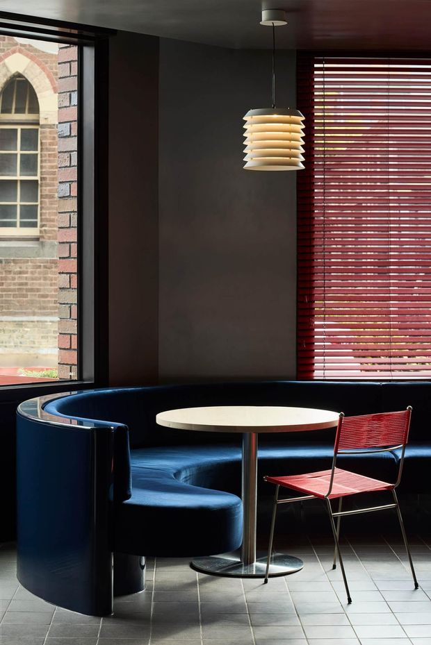 A compact, dark-toned interior seating area with a curved built-in navy banquette wrapping around a small round pedestal table and a red woven metal chair. The space has large black-framed windows, deep red horizontal blinds, dark walls, and a sculptural layered pendant light. Pale square floor tiles provide contrast, while an older brick-and-stone building is visible through the glazing. The composition has a refined mid-century-inspired, editorial character with strong geometric lines and dramatic natural light.