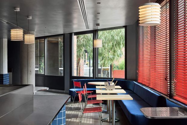 A modern commercial dining or café interior with a long navy upholstered banquette, small tables, red metal chairs, pendant lights, and large glazed openings. Dark painted walls and ceiling contrast with blue tiled joinery and extensive red horizontal blinds. A polished concrete or grey tile floor and exposed structural concrete column contribute to the industrial-contemporary character.