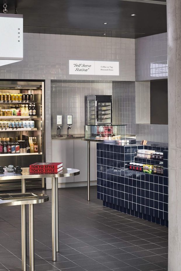 A compact self-serve café or specialty food retail interior with a dark tiled floor, pale square-tile walls, dark ceiling, stainless-steel display shelving, refrigerated beverage cases, beverage dispensers, and a navy-blue tiled service counter. Slim stainless-steel tables and shelving create a clean, highly considered commercial fit-out. Signage identifies a self-serve station with coffee and tea.