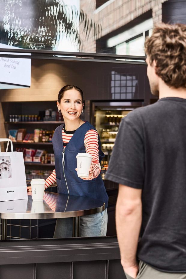 Photograph of a compact modern café or retail coffee counter viewed from the customer side. A barista stands behind a dark counter handing a takeaway cup to a customer, with shelves of packaged goods, a refrigerated display, dark tiled joinery, timber wall lining, and reflective glazing visible. The scene has a contemporary commercial fit-out with navy, black, timber, and stainless-steel details.