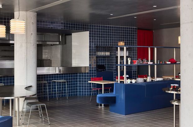 A contemporary commercial hospitality interior, likely a café or restaurant, with a dark blue tiled wall, stainless-steel kitchen and bar equipment, a large navy service counter with open shelving, integrated booth seating, café tables and stools. Concrete columns, dark ceilings, grid-pattern floor tiles, and vivid red wall accents create an industrial, graphic atmosphere.
