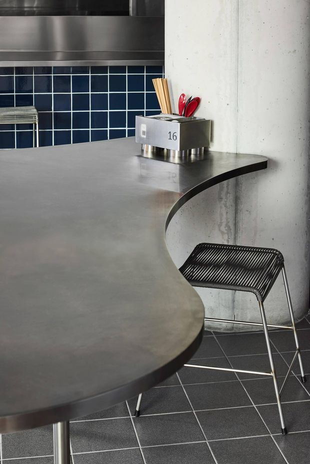 Close-up view of a sleek contemporary kitchen or commercial-style kitchenette with a large curved-edge grey concrete or stone island benchtop. The island features a stainless-steel utensil container, dark blue square tiled splashback, stainless-steel cabinetry or rangehood elements, and a black metal wire bar stool. A raw concrete wall and dark tiled floor reinforce the industrial character.