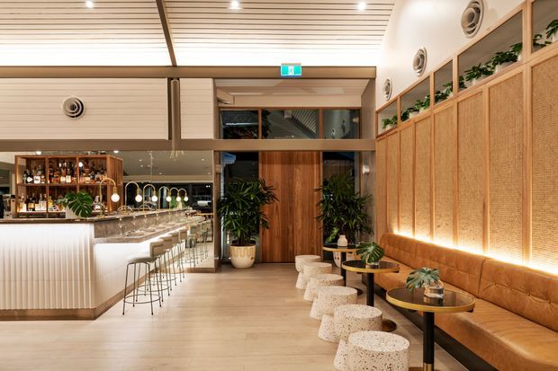 A refined contemporary hospitality interior, likely a restaurant or bar, featuring a long illuminated upholstered banquette, terrazzo stools and small round tables, a ribbed bar counter with stools, timber wall and ceiling finishes, woven-screen wall panels, integrated planter niches, and warm ambient lighting. The space has a high ceiling with linear slatted detailing and visible mechanical ventilation outlets.