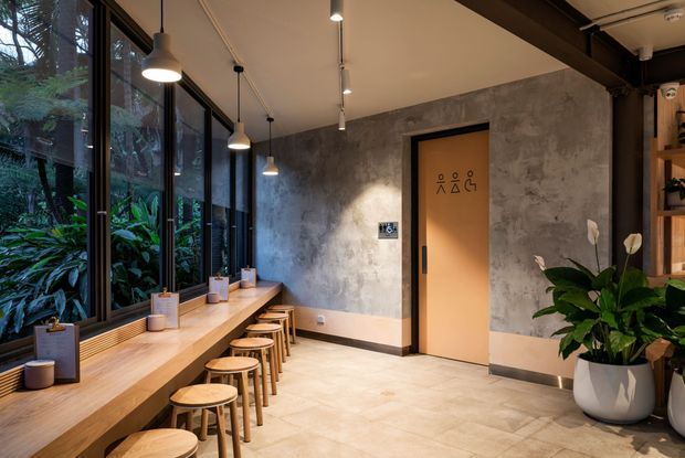 A contemporary commercial interior lounge or waiting area with a long timber window seat and counter lined with stools, floor-to-ceiling glazing overlooking lush greenery, exposed dark structural beams, concrete-look walls, pendant lighting, and a restroom door. Built-in shelving and large potted plants add warmth to the industrial-inspired space.