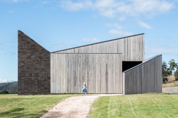 Exterior view of a contemporary, sculptural single-storey building with steeply angled rooflines and façades clad in vertical weathered timber. A dark textured brick wall forms one end, while a gravel path crosses the lawn toward a large central timber door. The rural setting and clear blue sky are visible around the structure.