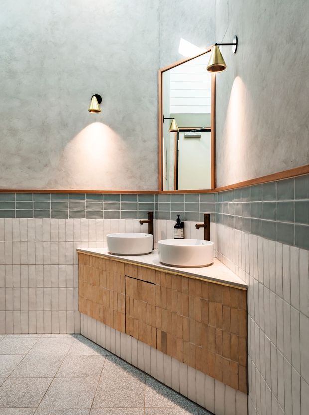 A contemporary bathroom with a double vessel-sink vanity, textured timber cabinetry, pale stone benchtop, and wall-mounted brass taps. The walls combine grey-green glazed rectangular tiles with light vertical tiles and a textured grey plaster upper wall. A large timber-framed mirror and warm brass wall sconces add contrast, while large grey floor tiles complete the space.