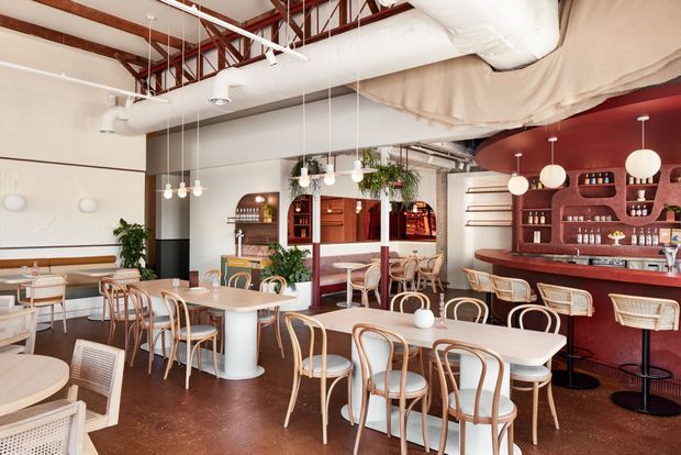 A bright contemporary café or restaurant interior with a visible bar, dining tables, bentwood chairs, banquette seating, pendant lights, abundant plants, and exposed services overhead. The space combines warm timber joinery with a distinctive deep red bar wall and polished dark flooring.