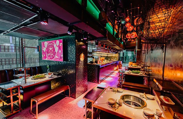A vibrant, dramatic Asian-inspired restaurant interior with multiple dining tables, built-in grill tables, banquette seating, a long service counter, and an open kitchen or preparation area. The space features dark walls and ceilings, exposed black structural and services elements, reflective metallic surfaces, vivid pink and green accent lighting, and numerous warm pendant lights. Large glazing is visible along one side, while patterned wall panels and a bright digital artwork add visual interest.
