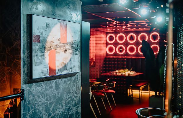 A dark, atmospheric commercial hospitality interior with a richly textured feature wall and framed abstract artwork in the foreground. Through the opening is a bar or lounge area with tables and chairs, dramatic red neon ring lighting, reflective ceiling details, and a strong red-and-black palette.