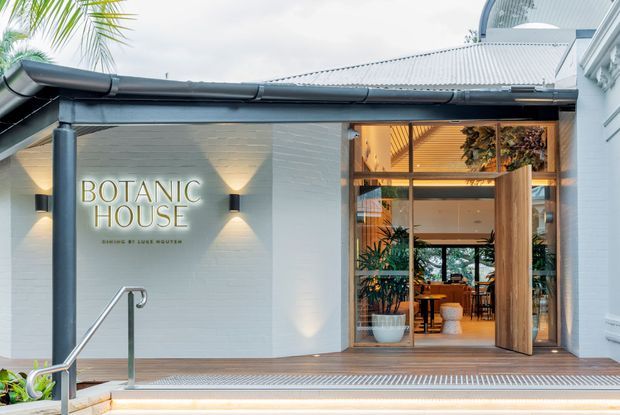 Exterior entrance to Botanic House, featuring a light-painted masonry facade, corrugated metal roof, dark metal gutters and posts, timber decking, broad steps with a stainless-steel handrail, and large glazed timber-framed doors opening into a warmly lit, plant-filled interior. A curved translucent roof element is visible above the entry.
