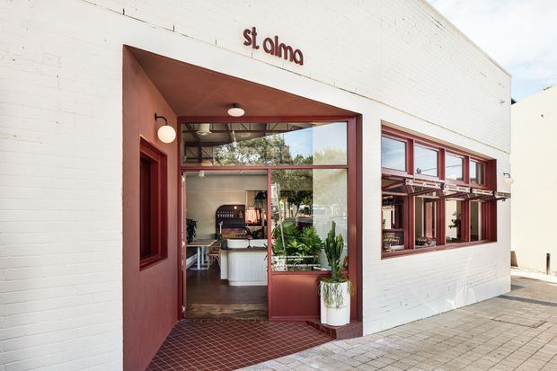Exterior view of a small commercial venue labelled “St. Alma,” with a white-painted brick facade, deep red entry recess and window frames, large glazed openings, and a tiled entrance threshold. The interior is partially visible through the open doorway and windows, showing a counter, pendant lighting, plants, and seating or dining fixtures.