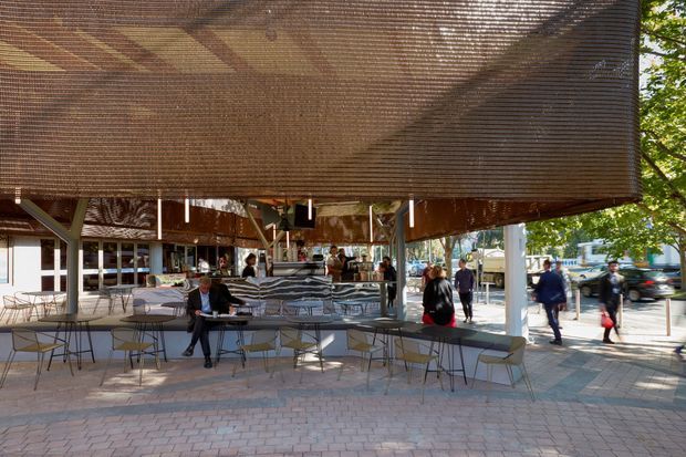 Open-air commercial dining and hospitality space beneath a large suspended timber slatted canopy. The covered terrace contains numerous tables and chairs, a long counter or service area, and people dining or walking through. The space opens onto a paved urban pedestrian area with trees and surrounding buildings visible beyond.