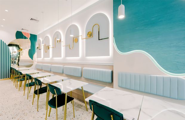 Bright contemporary commercial dining space with a long banquette, multiple white tables, and teal upholstered chairs. The interior features an aqua and white sculptural wall treatment with arched recessed niches, integrated curved lighting, gold decorative accents, pendant lights, and a terrazzo-style floor. The palette is fresh, playful, and refined, resembling a modern café or restaurant.