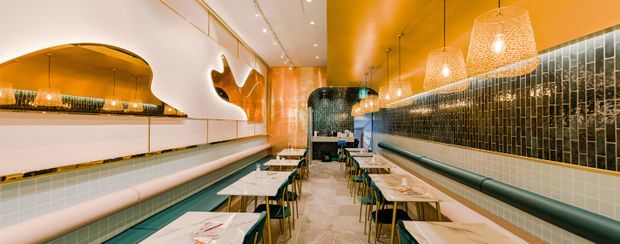 A narrow, elongated contemporary commercial dining space, likely a restaurant or café, viewed down the central aisle. It features rows of small tables with green chairs, continuous dark green upholstered banquette seating, glossy green tiled walls, pale tiled flooring, a warm orange ceiling and walls, sculptural organic wall openings, and woven pendant lights. The space has a polished, playful, retro-inspired atmosphere with strong linear perspective.