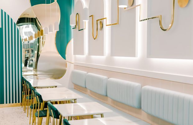 A stylized contemporary commercial dining or hospitality interior with a long banquette upholstered in pale blue, small white marble-look tables, and teal dining chairs with slender gold-toned legs. The space features sculptural pendant lights, curved mirrored forms, teal vertical wall striping, white wall panels with integrated lighting, and decorative gold linear details. The palette is bright with teal, white, pale blue, and gold accents.