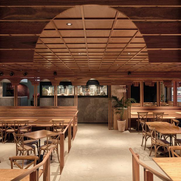 A warmly lit restaurant or hospitality dining space with extensive dark timber wall and ceiling finishes. The ceiling features a large central arched area with a decorative timber lattice or woven-panel grid and recessed downlights. Timber-framed dining tables and chairs fill the room, while a central bar or service counter with dark stone or mottled panels sits at the rear, backed by glazed display areas and mirrors. Indoor plants and arched openings add visual detail. The atmosphere is refined, warm, and inviting.