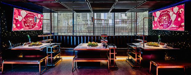 A dark, high-energy commercial dining or entertainment venue with multiple rectangular dining tables, long upholstered banquette seating, and exposed metal ceiling grid. Black walls feature small star-like points, while vivid red illuminated wall screens and blue, red, and purple accent lighting create a theatrical atmosphere. Large windows line the rear of the space, and the furniture uses dark timber or laminate surfaces with black and metallic detailing.
