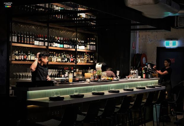 A dimly lit contemporary bar and dining counter, with extensive open bottle shelving behind the bar, a long illuminated counter, bar stools, glassware, and bartenders preparing drinks. The space has dark finishes, exposed services, and atmospheric green-tinted under-counter lighting.