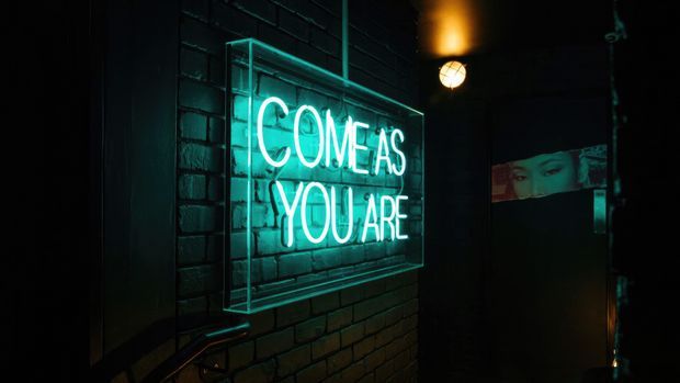 Dark, moody interior entrance or corridor with a glowing turquoise neon sign reading “COME AS YOU ARE” mounted on a black painted brick wall. A warm overhead wall light and a partially visible graphic or reflection appear in the background, creating a nightclub or bar-like atmosphere.