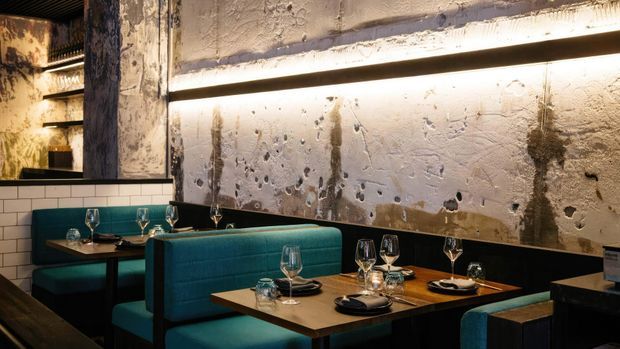 Industrial-style restaurant dining area with teal upholstered booth seating, dark wooden tables, exposed distressed concrete walls, black trim, and warm linear lighting. Table settings with wine glasses and plates create an intimate, refined atmosphere.