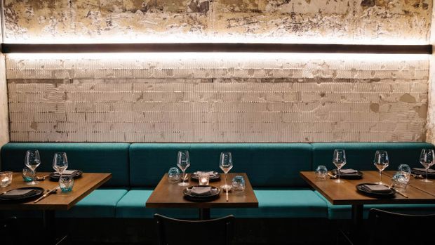 A dimly lit contemporary restaurant dining area with teal upholstered banquette seating arranged around dark timber tables. The tables are set with plates, wine glasses and glassware, while a textured exposed masonry wall and a long linear wall light create an industrial, refined atmosphere.