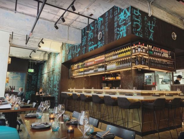 Interior view of a contemporary restaurant and bar, featuring a long illuminated drinks bar with extensive bottle shelving, dark patterned cabinetry, turquoise handwritten-style wall graphics, counter seating, and dining tables set with glassware and tableware. The ceiling has exposed services and track lighting, giving the space an industrial character.