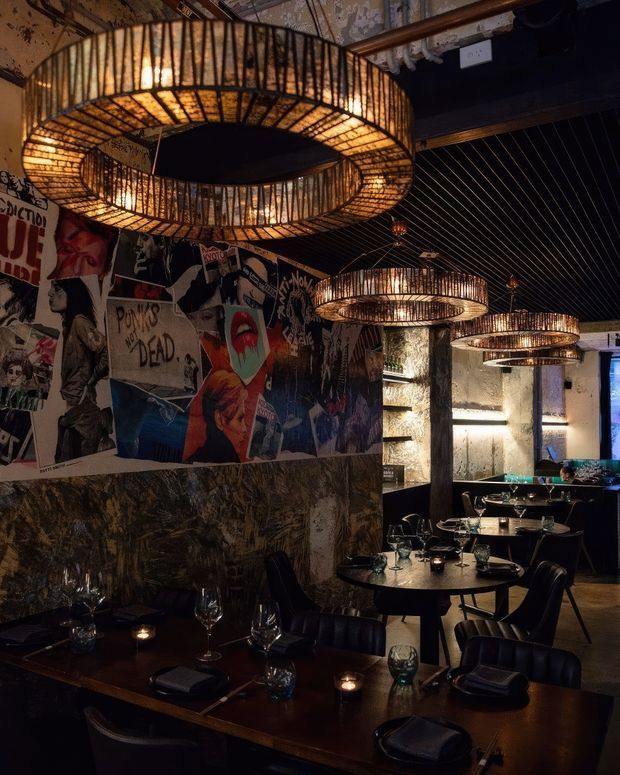 A moody, darkly atmospheric restaurant or bar interior with multiple large circular woven pendant lights, black slatted ceiling, textured stone or concrete columns, dark dining tables and upholstered chairs. The walls feature a bold collage of punk and pop-art posters, creating an industrial, eclectic dining setting.