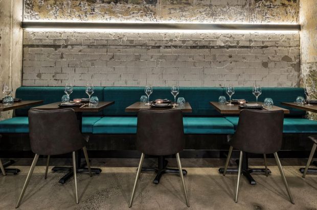 A moody contemporary restaurant dining area with a long teal upholstered banquette, dark dining tables, and matching dark leather chairs. Exposed rough concrete or plaster wall surfaces and a narrow linear ceiling light create an industrial, textural atmosphere. Tables are set with wine glasses and place settings.