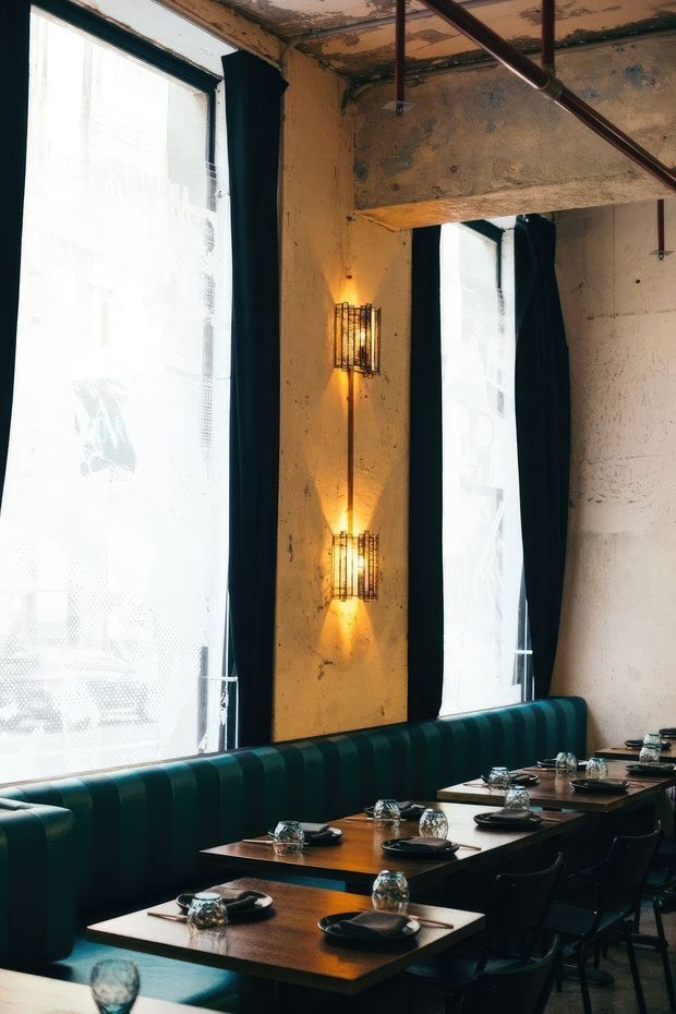 A moody industrial-style restaurant dining area with tall windows, dark curtains, exposed aged concrete walls and ceiling structure, caged wall sconces, and a long deep teal upholstered banquette. Dark timber dining tables are set with black plates, glassware, and place settings, paired with dark chairs.