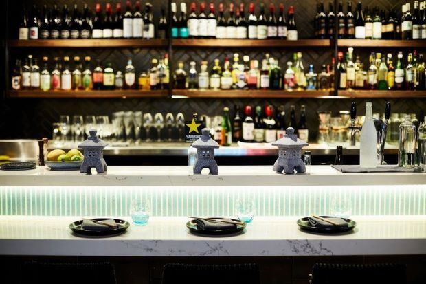 A dimly lit bar or restaurant counter with extensive backbar bottle shelving, hanging glassware, a bright illuminated tiled front, and three place settings arranged along the counter. Decorative stone-like objects and bar equipment sit on the countertop.