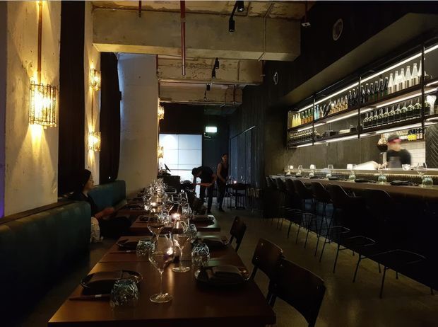 A dimly lit, industrial-style restaurant interior with a long dining table set with glassware and tableware, dark upholstered banquette seating along the left wall, and a long bar with stools and illuminated bottle shelving on the right. Exposed concrete, dark ceilings, pendant and wall lighting, and a visible service counter create a moody hospitality setting.