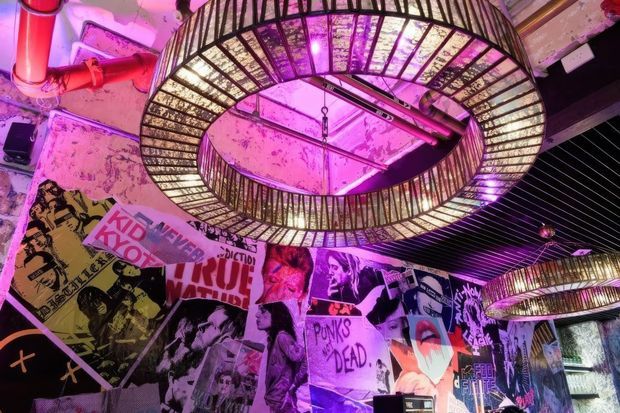 A dramatic, heavily decorated interior with exposed, distressed surfaces and large circular metal-and-glass pendant lights suspended from the ceiling. The walls are covered with layered punk and street-art posters, while vivid magenta and purple lighting creates an energetic nightlife atmosphere. Exposed red-painted pipework and dark ceiling elements contribute to an industrial, alternative venue aesthetic.
