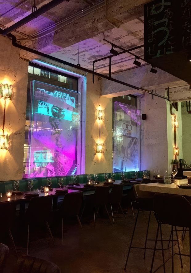 A dimly lit contemporary restaurant or bar interior with exposed, weathered concrete ceilings and walls, suspended black track lighting, and large windows displaying vivid purple and turquoise projections. Tables are set with glassware and candles, with dark dining chairs and bar stools creating an atmospheric, industrial dining environment.