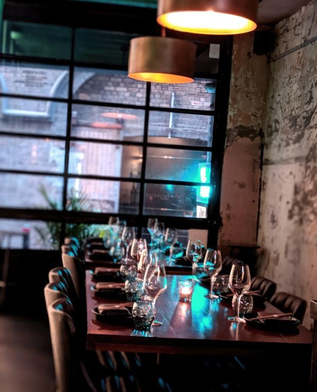 A moody restaurant dining space with a long dark timber communal table, place settings, wine glasses, upholstered chairs, exposed textured masonry walls, large black-framed industrial-style windows, and warm copper pendant lights. Blue and turquoise light reflections create a dramatic atmospheric contrast.