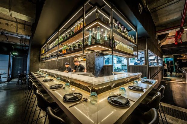 A dramatic, contemporary commercial restaurant interior centred on a large angular island bar and counter. Suspended illuminated glass-and-metal shelving displays bottles and glassware above the bar, while a U-shaped dining counter is set with dark plates, cutlery and glasses. Open kitchen activity is visible behind the bar, with dark industrial ceiling and wall finishes, exposed services, and warm integrated lighting.
