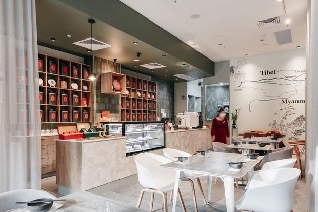 Bright contemporary commercial café interior with a service counter, refrigerated display case, espresso station, extensive dark timber shelving displaying packaged products, and a dining area furnished with white chairs and tables. A large illustrated wall graphic forms a feature backdrop, while a dark painted ceiling soffit contrasts with pale timber-look counter finishes and patterned wall surfaces.
